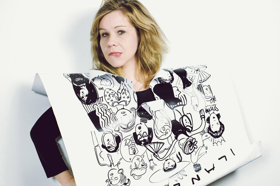 Na zdjęciu kobieta o blond włosach, jej kark i tułów pokrywa kartka papieru z rysunkami.