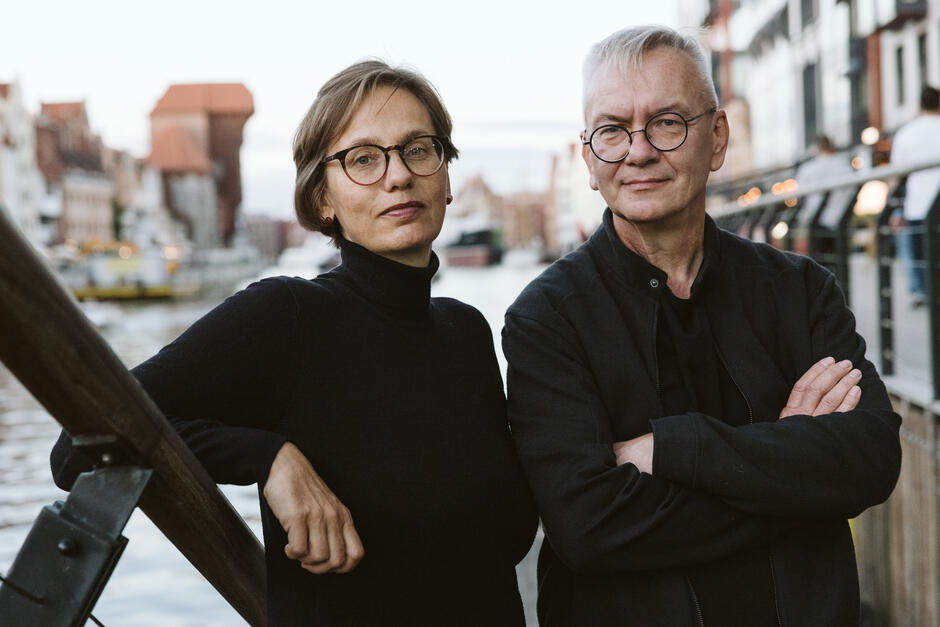 Na zdjęciu kobieta i mężczyzna w czarnych strojach, oboje w okularach, w tle zabytkowe budynki Gdańska.