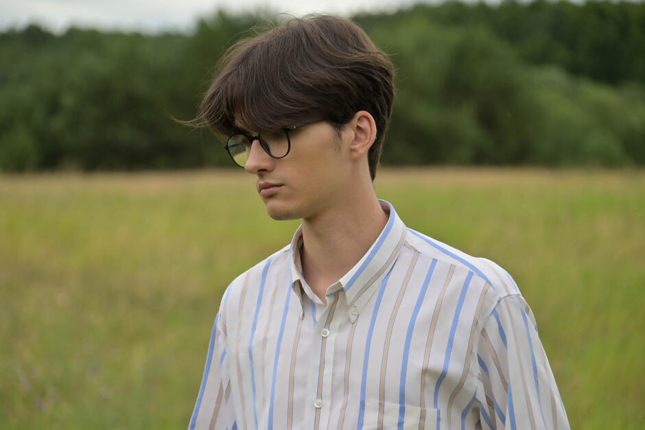Na zdjęciu młody mężczyzna w okularach korekcyjnych, w koszuli w paski na tle łąki i drzew.