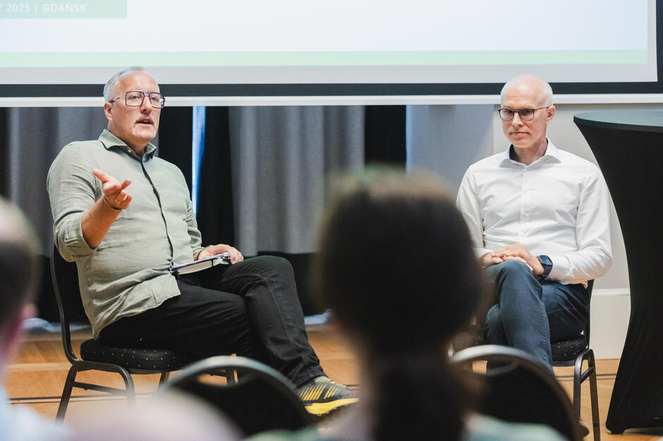 Na zdjęciu dwóch mężczyzn bierze udział w spotkaniu lub panelu dyskusyjnym, siedząc na krzesłach przed publicznością. Jeden z nich gestykuluje i trzyma notes, co sugeruje, że właśnie zabiera głos lub odpowiada na pytanie.