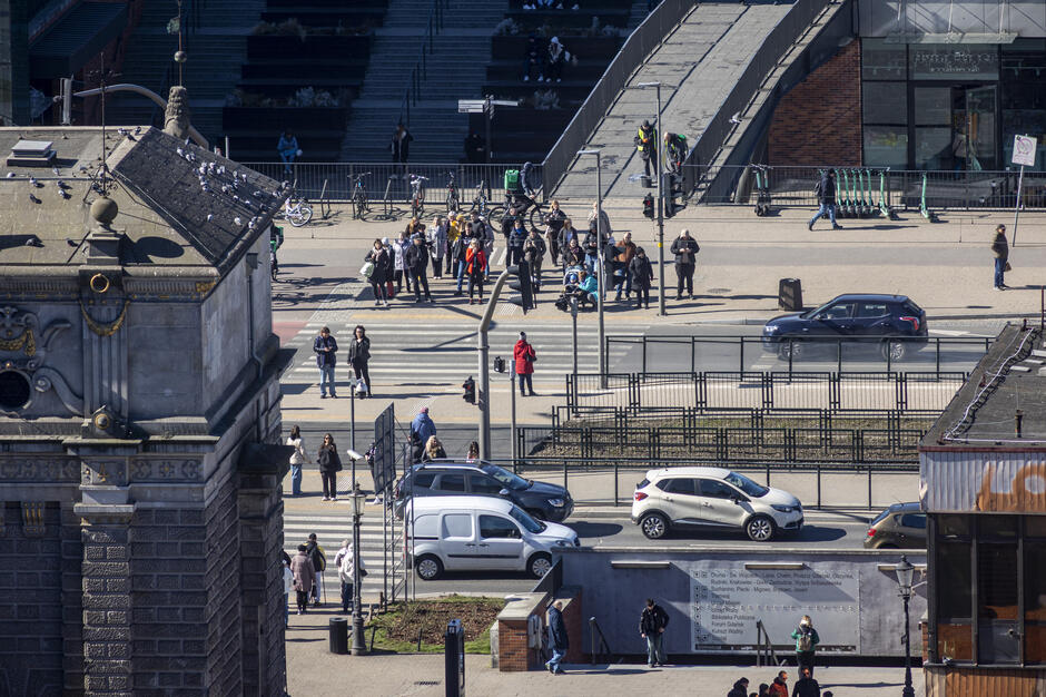 Zdjęcie przedstawia ruchliwe przejście dla pieszych w centrum miasta, gdzie duża grupa ludzi czeka na zmianę świateł, a część osób już przechodzi przez jezdnię. W tle widać nowoczesne schody i rampę, zaparkowane hulajnogi, a z przodu fragment zabytkowego budynku i jadące samochody.