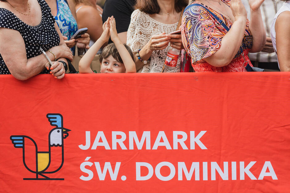 Zdjęcie przedstawia tłum ludzi stojących za czerwoną barierką z napisem „Jarmark św. Dominika” oraz grafiką stylizowanego koguta. W centrum kadru uśmiechnięty chłopiec z uniesionymi rękami wygląda zza baneru, otoczony przez dorosłych trzymających telefony i napoje.