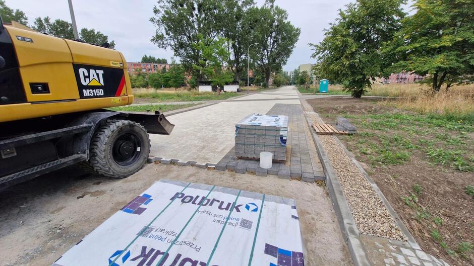 na zdjęciu z lewej fragment żółtej koparki, obok sztapelek nowej kostki betonowej, widać też układaną z kostki jezdnię