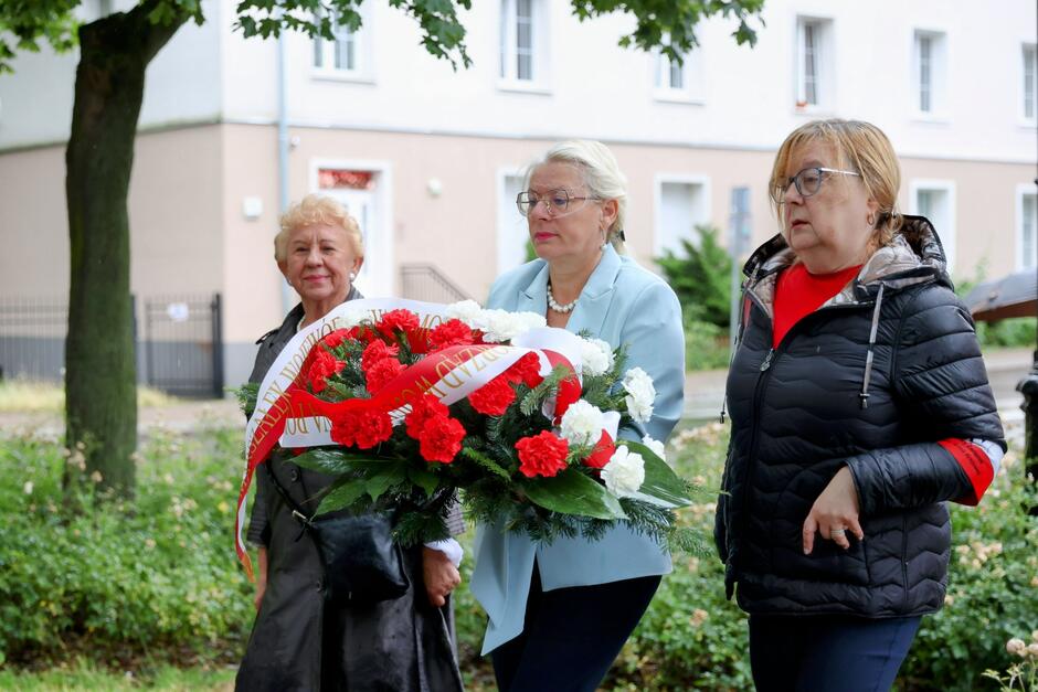 Na zdjęciu widzimy trzy kobiety biorące udział w uroczystości upamiętniającej. Centralna postać niesie okazały wieniec z biało-czerwonych goździków, przewiązany biało-czerwoną wstęgą z napisem – prawdopodobnie odnoszącym się do bohaterów walki o niepodległość Polski. Pozostałe dwie kobiety idą obok, towarzysząc jej w tej symbolicznej chwili. Uroczystość odbywa się w przestrzeni miejskiej, przy budynkach mieszkalnych. Wszystkie trzy kobiety ubrane są w sposób stosowny do powagi wydarzenia – elegancko i z szacunkiem. Jedna z nich ma opaskę na rękawie, co może sugerować przynależność do środowiska kombatanckiego lub patriotycznego.