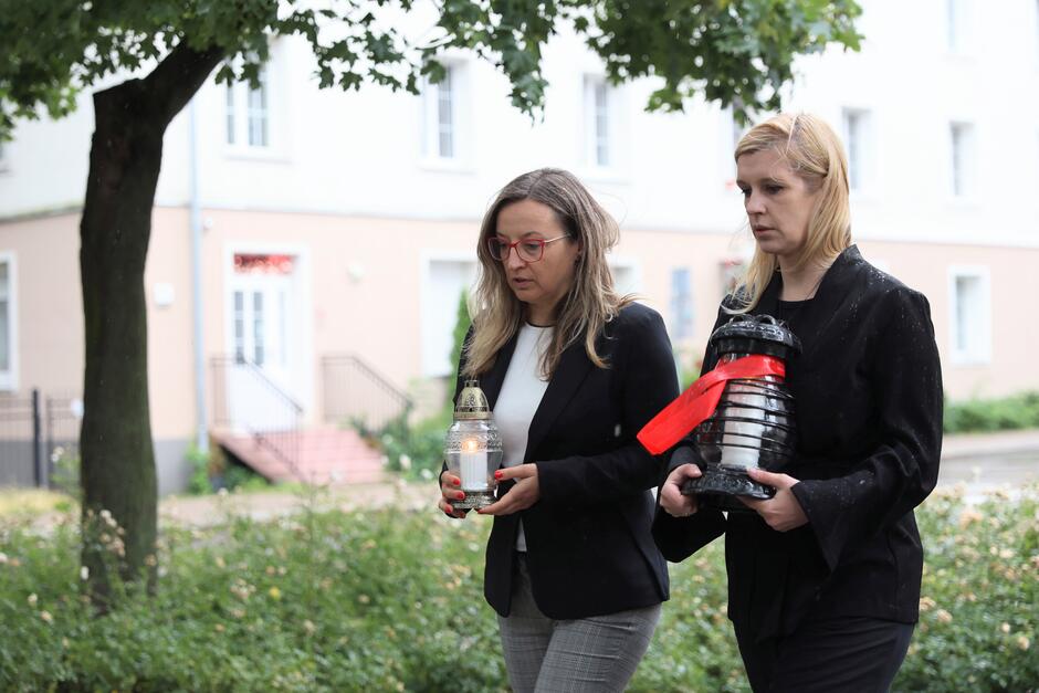 Na zdjęciu widzimy dwie kobiety niosące znicze – jedna z nich trzyma biały znicz, druga czarny znicz przewiązany czerwoną wstążką. Obie panie mają poważne, skupione wyrazy twarzy, co świadczy o uroczystym i podniosłym charakterze wydarzenia. Najprawdopodobniej zmierzają w stronę miejsca pamięci, by złożyć znicze jako symbol szacunku i pamięci o zmarłych. Tło i otoczenie są spójne z wcześniejszymi zdjęciami – miejskie osiedle, drzewa, krzewy i budynki mieszkalne. To wszystko wskazuje, że zdjęcie zostało wykonane w tym samym miejscu, w czasie uroczystości upamiętniającej – być może rocznicę Powstania Warszawskiego, Dzień Pamięci Ofiar czy innego wydarzenia związanego z historią Polski.