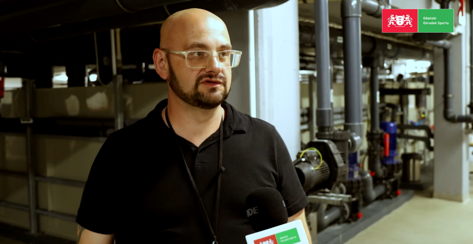 Zdjęcie przedstawia mężczyznę udzielającego wywiadu w pomieszczeniu technicznym, prawdopodobnie na zapleczu basenu – w tle widoczne są rury, pompy i instalacje. Mężczyzna trzyma mikrofon z logo Gdańskiego Ośrodka Sportu, co sugeruje, że wypowiada się jako pracownik lub ekspert.