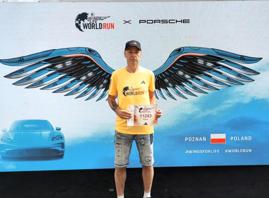 Zdjęcie przedstawia uczestnika biegu Wings for Life World Run w Poznaniu, ubranego w żółtą koszulkę z logo wydarzenia i trzymającego numer startowy 11243. W tle znajduje się efektowna grafika z mechanicznymi skrzydłami, logotypami Wings for Life i Porsche oraz flagą Polski.