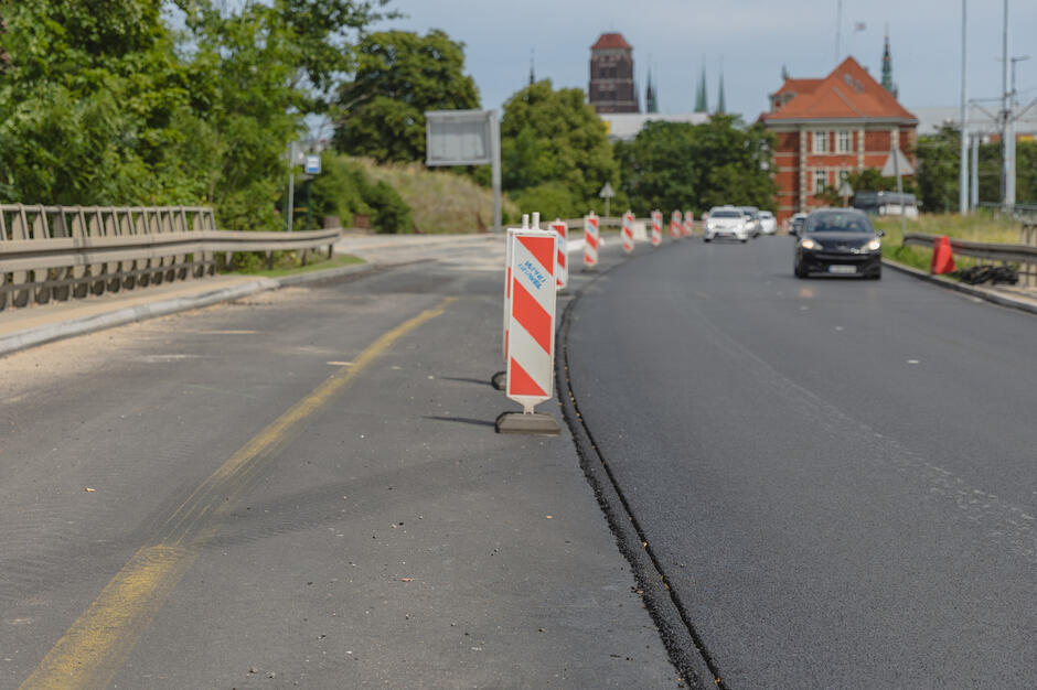 na zdjęciu jezdnia, po prawej widać nową nawierzchnię, na jezdni ustawione są też znaki drogowe, po prawej jadą samochody