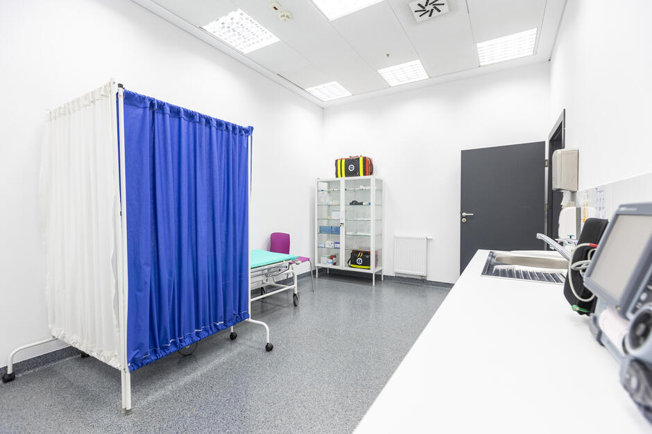 Na zdjęciu widać jasny, sterylny gabinet medyczny z łóżkiem do badań osłoniętym niebieską zasłoną oraz regałem z wyposażeniem ratowniczym. Na blacie znajduje się sprzęt medyczny, a w rogu sali stoi otwarte drzwi. 