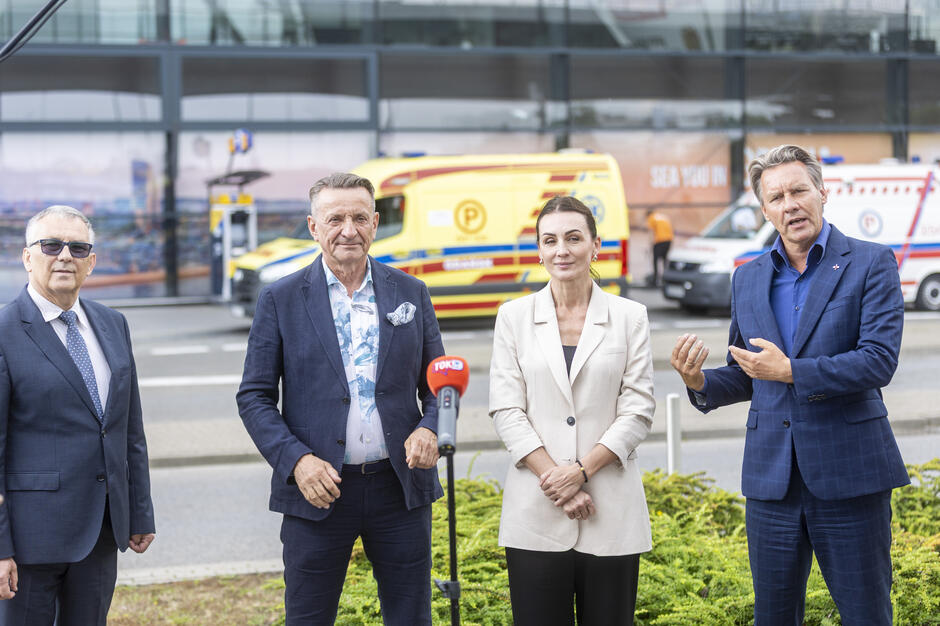 Na zdjęciu cztery osoby w eleganckich strojach stoją przed mikrofonem podczas konferencji prasowej, w tle widoczne są karetki pogotowia. Wydarzenie odbywa się przed przeszklonym budynkiem, a atmosfera sugeruje oficjalne wystąpienie lub prezentację. 