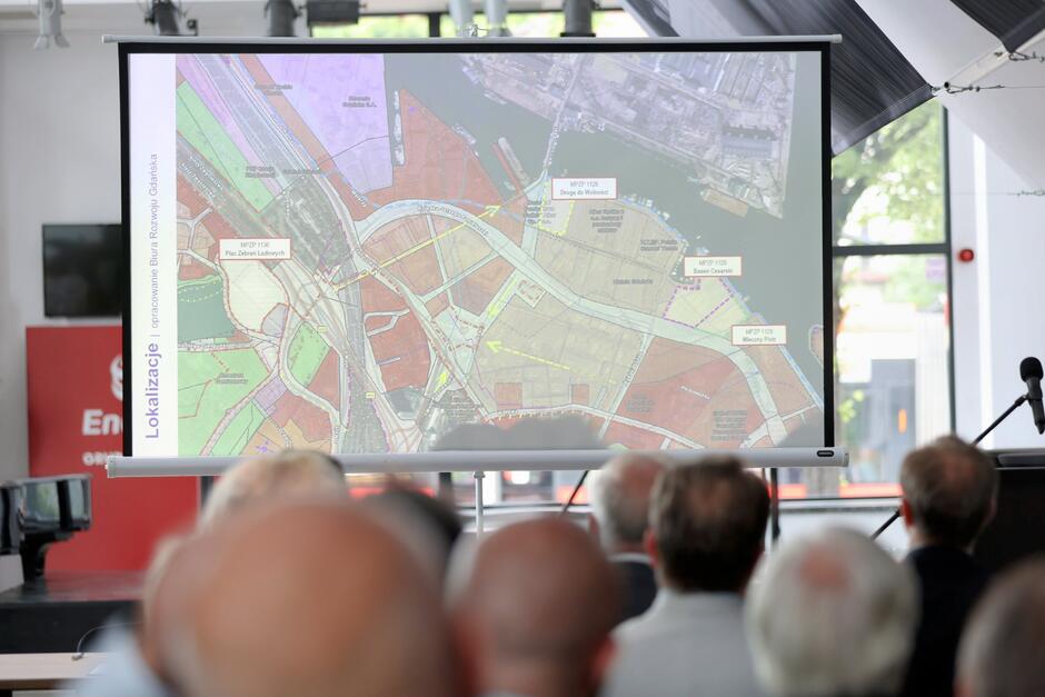 Na ekranie widoczna jest szczegółowa mapa urbanistyczna z oznaczeniem planowanych terenów i dróg w Gdańsku, prezentowana podczas oficjalnego spotkania. W tle widać publiczność śledzącą prezentację oraz ustawiony mikrofon.