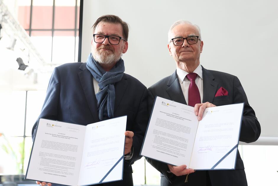 Dwaj mężczyźni w garniturach pozują z dokumentami podpisanymi podczas oficjalnej uroczystości. Ich postawa i uśmiechy sugerują zakończenie ważnego etapu współpracy lub zawarcie umowy.