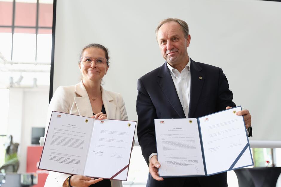 Kobieta i mężczyzna stoją razem, trzymając przed sobą podpisane dokumenty oznaczone herbami Gdańska i Województwa Pomorskiego. Oboje uśmiechają się, co świadczy o pozytywnym charakterze zawartego porozumienia