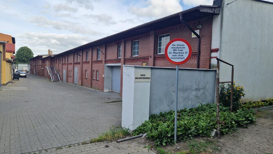 Znak zakazu ruchu, murek z tablicą Zaułek Jana Kwiatkowskiego, a za nim długi jednopiętrowy ceglany budynek