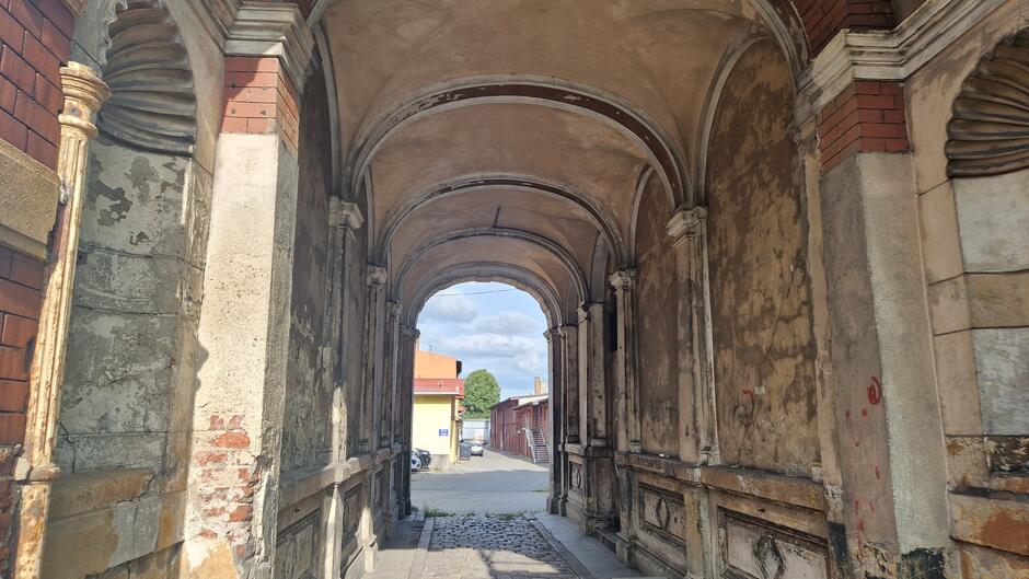 Z lewej strony bramy stoi wysoki metalowy słup wtopiony w spękaną ścianę kamienicy