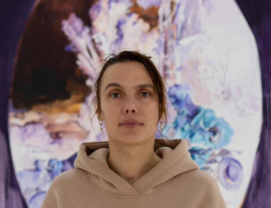 Na zdjęciu kobieta w jasnej bluzie, stojąca na tle obrazu.