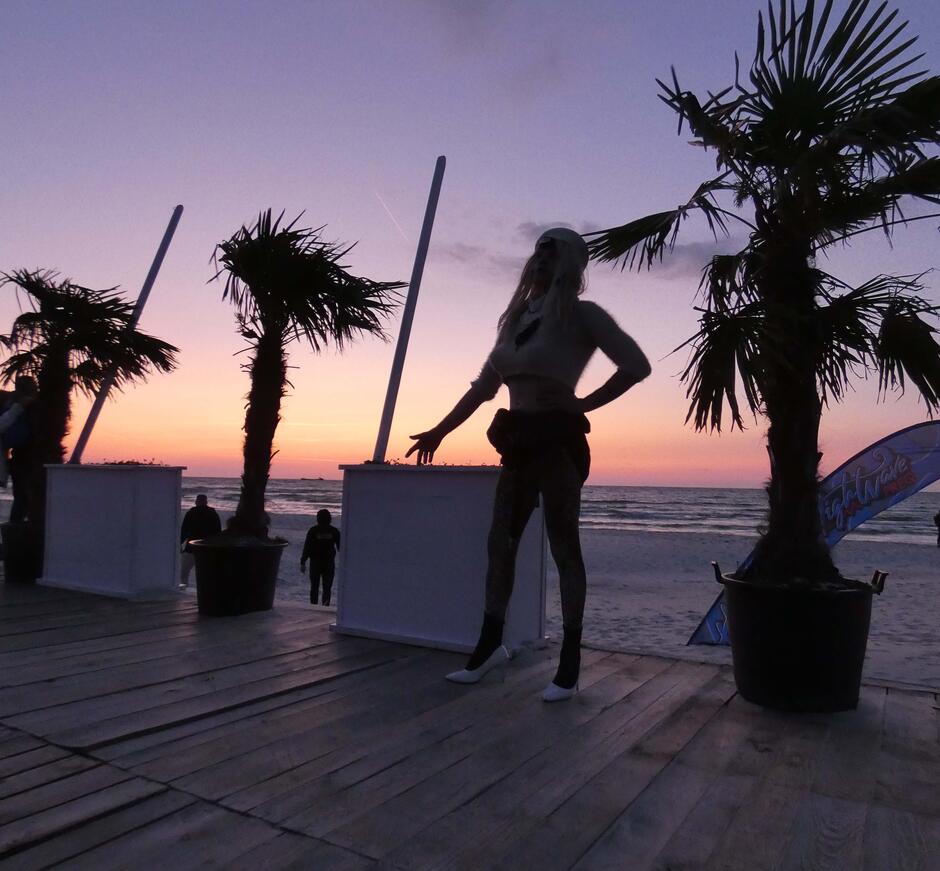 Na zdjęciu mężczyzna ubrany w strój kobiety, wśród palm, z widokiem na morze.