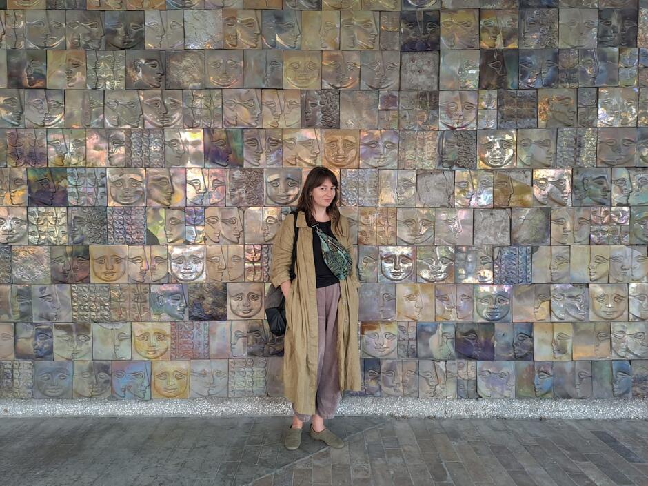 Na zdjęciu kobieta ubrana w jasnobrązowy płaszcz, stojąca na tle ściany z mozaiki