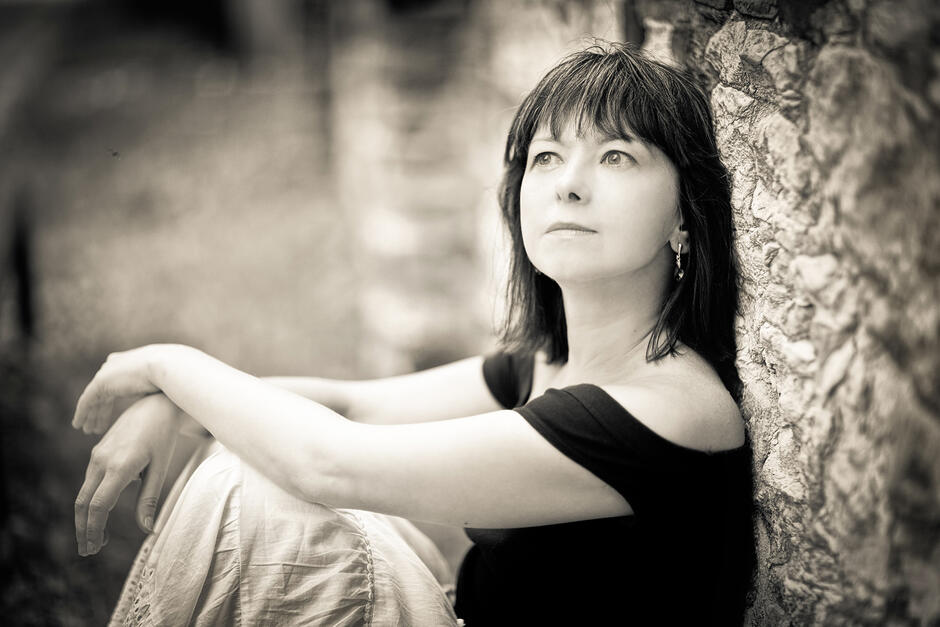 Na zdjęciu kobieta patrząca w lewo, oparta o mur, ubrana w czarną bluzkę oraz jasną spódnicę.