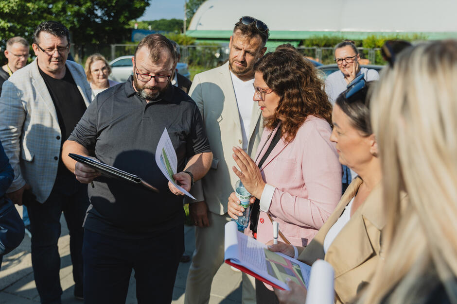 na zdjęciu grupa dorosłych osób stoi obok siebie, na pierwszym planie mężczyzna w średnim wieku w czarnej koszuli pokazuje coś na kartce kobiecie w różowej marynarce, także w średnim wieku