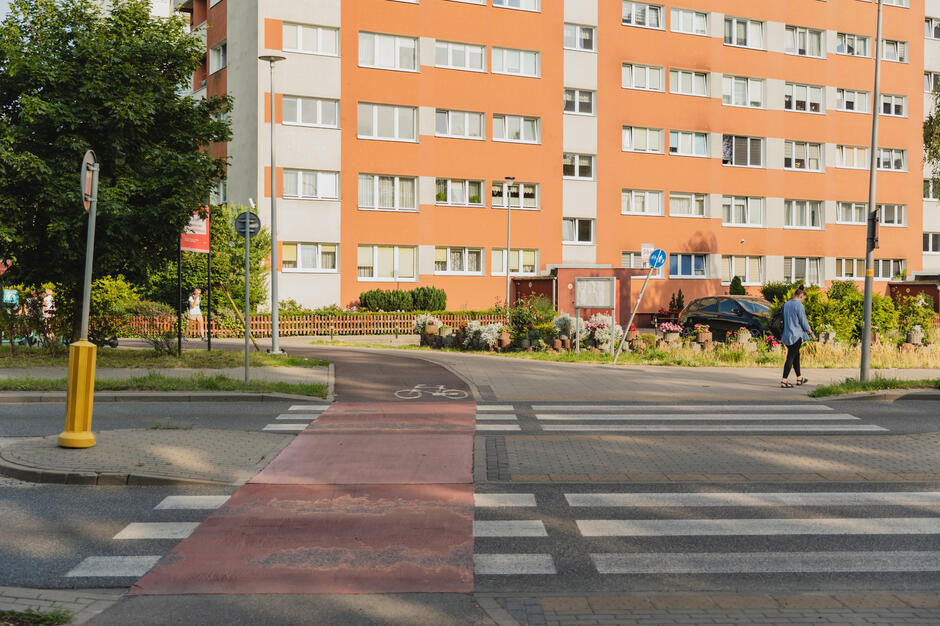 na zdjęciu przejście dla pieszych wraz z wyznaczonym przejazdem dla rowerów, w tle blok mieszkalny