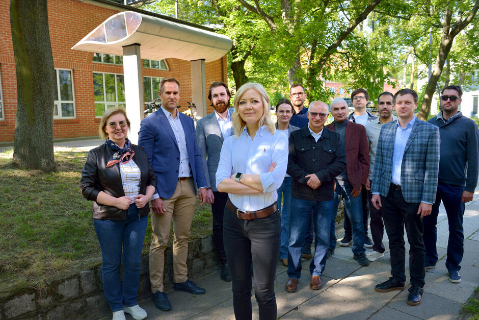 Na zdjęciu widać grupę kilkunastu osób stojących razem na tle budynku z czerwonej cegły, w otoczeniu zieleni. Na pierwszym planie stoi kobieta w białej koszuli z założonymi rękami, a reszta grupy pozuje za nią w swobodnej, ale profesjonalnej atmosferze.