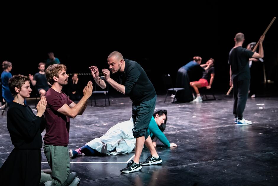 Na zdjęciu widzimy grupę aktorów podczas dynamicznej sceny teatralnej — niektórzy klęczą, inni gestykulują w napięciu, a na dalszym planie toczy się równoległa akcja. Całość rozgrywa się na pustej scenie, co podkreśla intensywność fizycznego i emocjonalnego wyrazu postaci