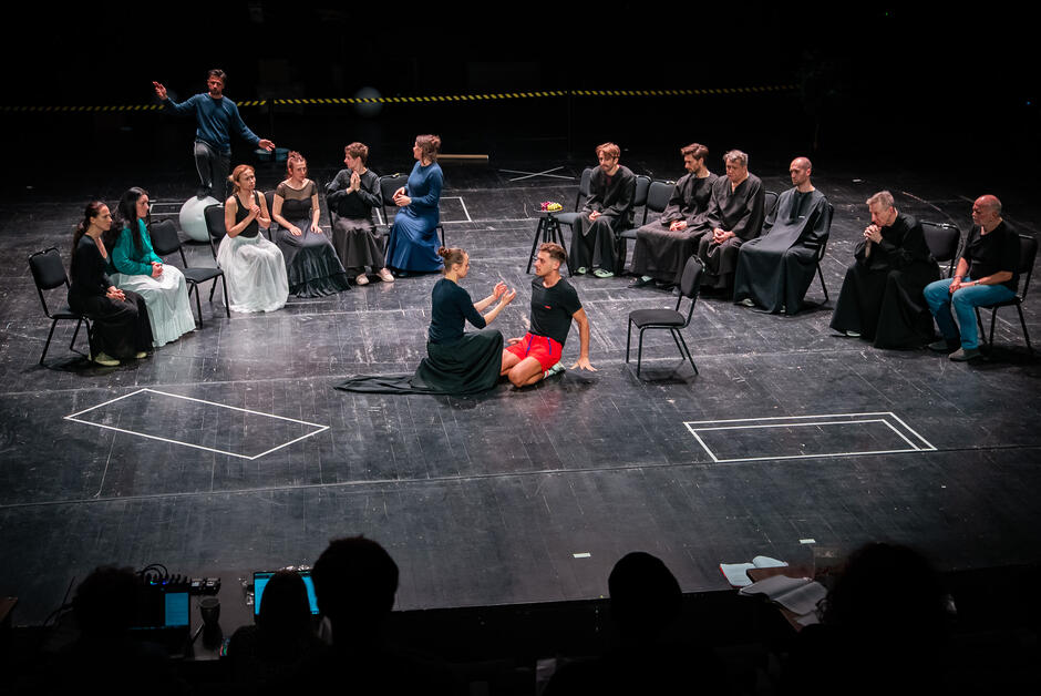 Zdjęcie ukazuje teatralną scenę zbiorową — aktorzy siedzą w półokręgu, a na środku para postaci prowadzi intensywną interakcję na siedząco i klęcząco. Kompozycja przypomina sądowy lub ceremonialny układ, a wyraziste gesty postaci sugerują dramatyczne napięcie.