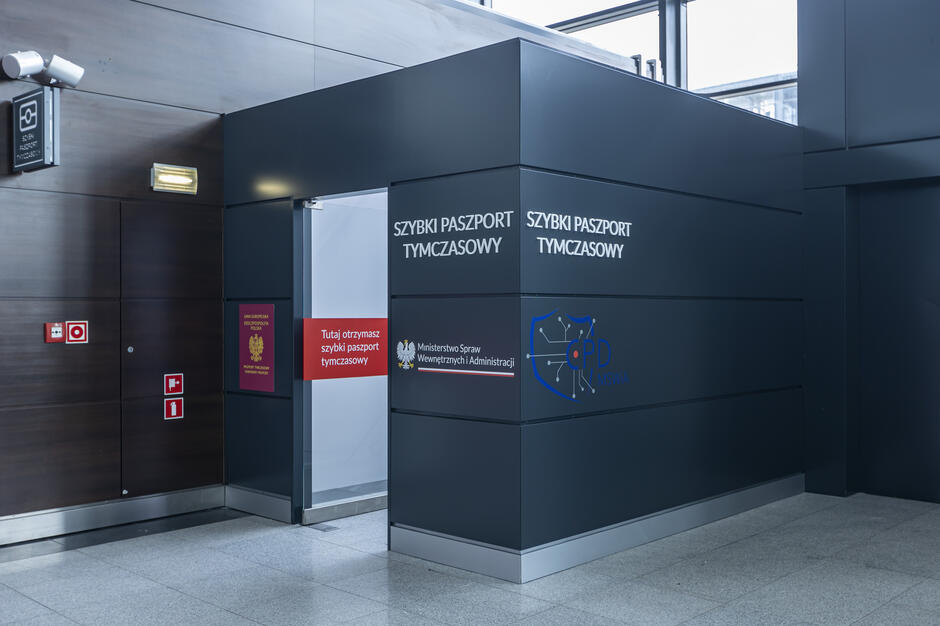 Na zdjęciu widoczny jest punkt wydawania szybkich paszportów tymczasowych, zlokalizowany na lotnisku i oznaczony logiem Ministerstwa Spraw Wewnętrznych i Administracji. Wejście do punktu jest wyraźnie oznaczone napisami informacyjnymi w języku polskim.