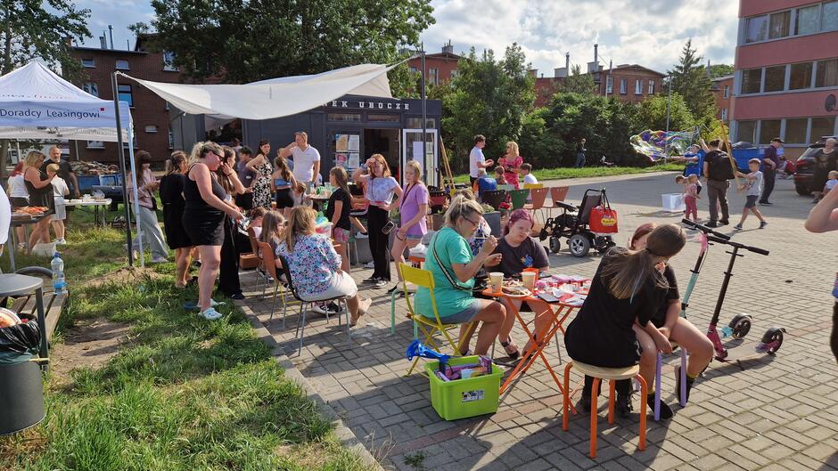 Na placu przed parterowym budynkiem z kontenerów siedzą ludzie przy stolikach, robiąc zmywalne tatuaże, doczepy do włosów, dalej stoły z jedzeniem - trwa festyn
