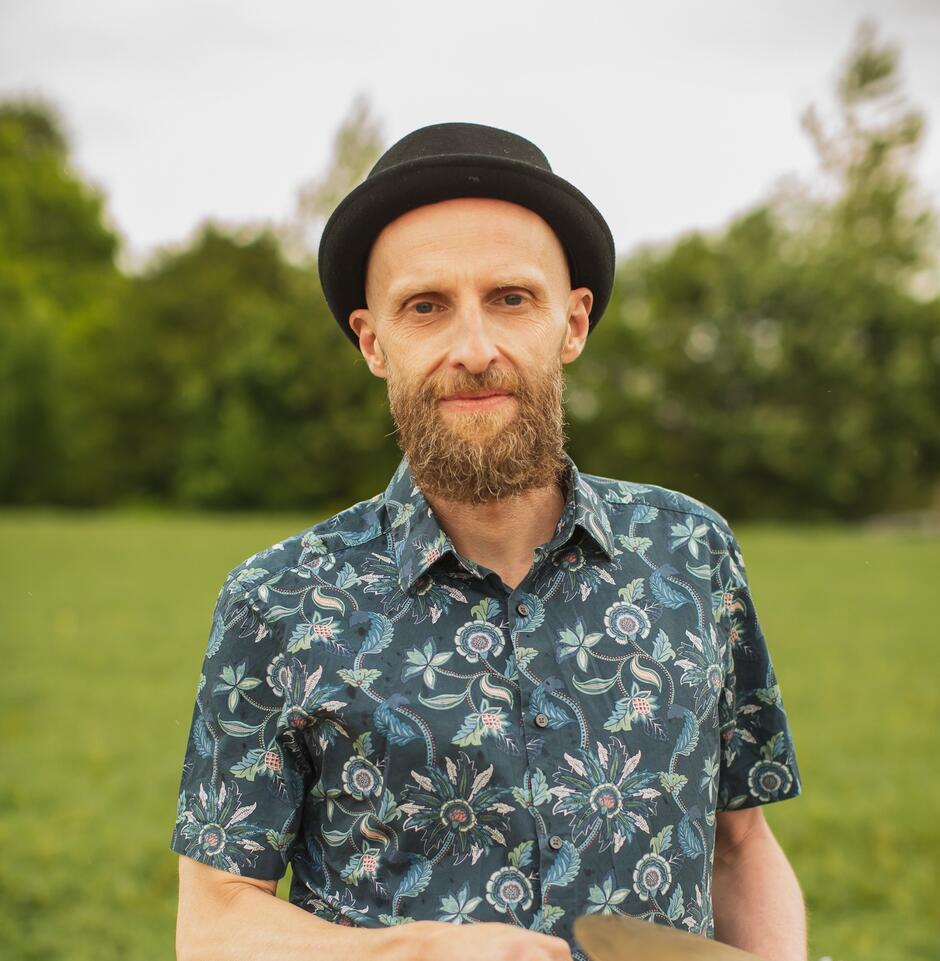 Na zdjęciu mężczyzna w czarnym kapeluszu i koszuli w kwiaty na tle zieleni.