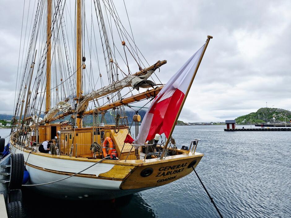 Na zdjęciu widzimy żaglowiec Generał Zaruski zacumowany przy nabrzeżu, z polską banderą powiewającą na rufie. W tle rozciąga się spokojny, norweski krajobraz z wyspami i niskimi chmurami na niebie.