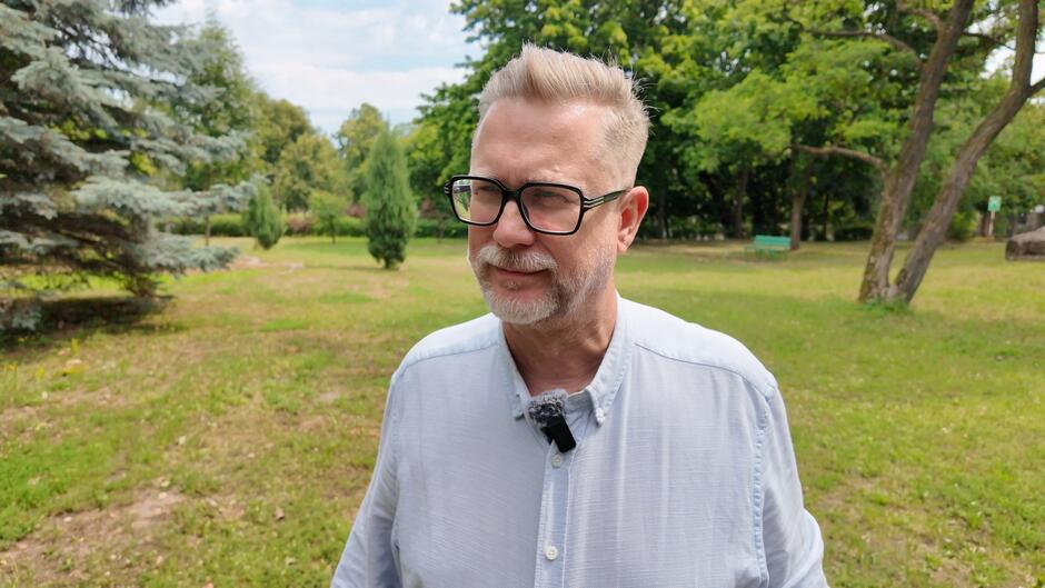 Na zdjęciu widzimy mężczyznę stojącego w parku lub ogrodzie. Ma krótkie, jasne włosy uczesane do góry i nosi ciemne okulary w grubych oprawkach. Ma również siwą brodę i wąsy. Ubrany jest w jasną koszulę z kołnierzykiem, a przy niej przypięty ma mikrofon krawatowy. W tle znajduje się zielona trawa, drzewa oraz parkowa ławeczka — otoczenie wygląda na spokojne i letnie.