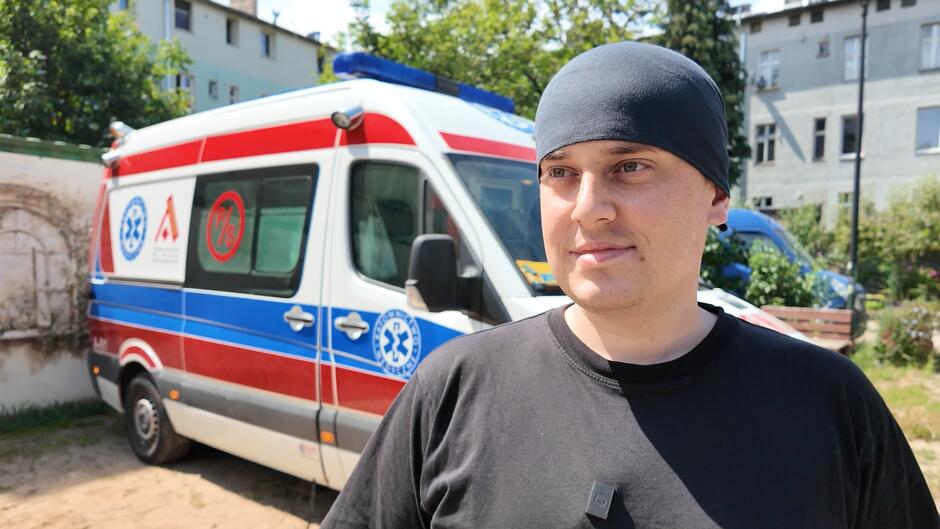 Na tym zdjęciu widać mężczyznę stojącego na tle karetki pogotowia. Ma na sobie czarną koszulkę i czarną opaskę lub chustę na głowie. Do koszulki przypięty ma mikrofon krawatowy. Mężczyzna patrzy lekko w bok i uśmiecha się delikatnie. W tle znajduje się ambulans z charakterystycznym czerwono-niebieskim pasem i emblematami ratownictwa medycznego, a także budynki mieszkalne oraz zieleń. Zdjęcie zostało wykonane w słoneczny dzień.