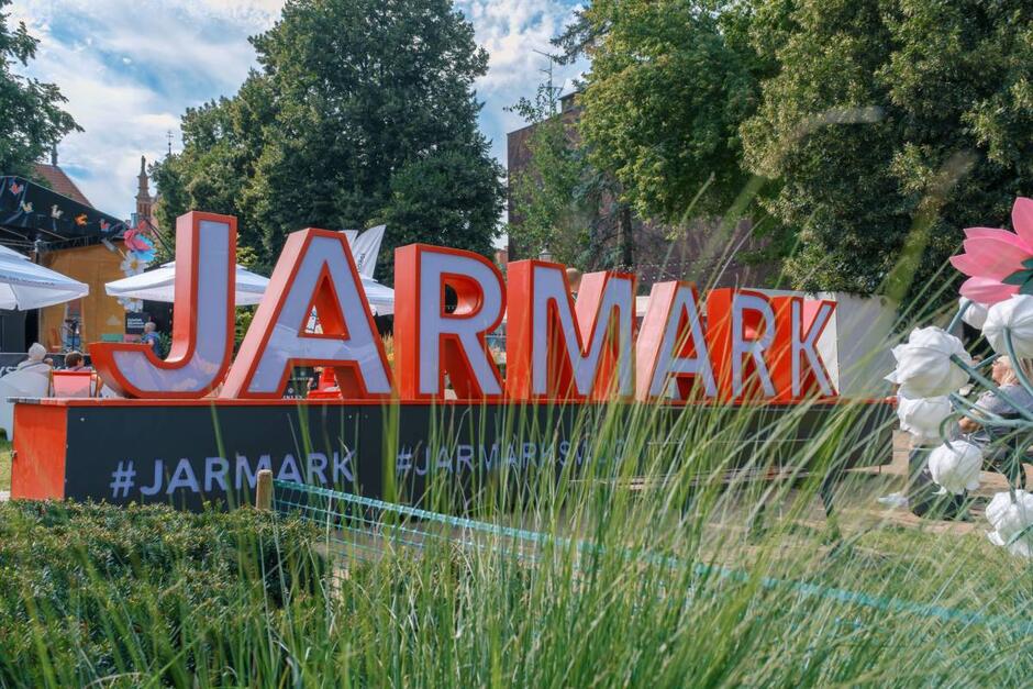 Duży czerwony napis „JARMARK” ustawiony w przestrzeni miejskiej wśród zieleni i trawy. W tle widoczne są stoiska z białymi parasolami, scena ozdobiona kolorowymi dekoracjami oraz ludzie odpoczywający w cieniu drzew. Na dolnej części instalacji widnieją hashtagi: #JARMARK, #JARMARKŚWDOMINIKA.