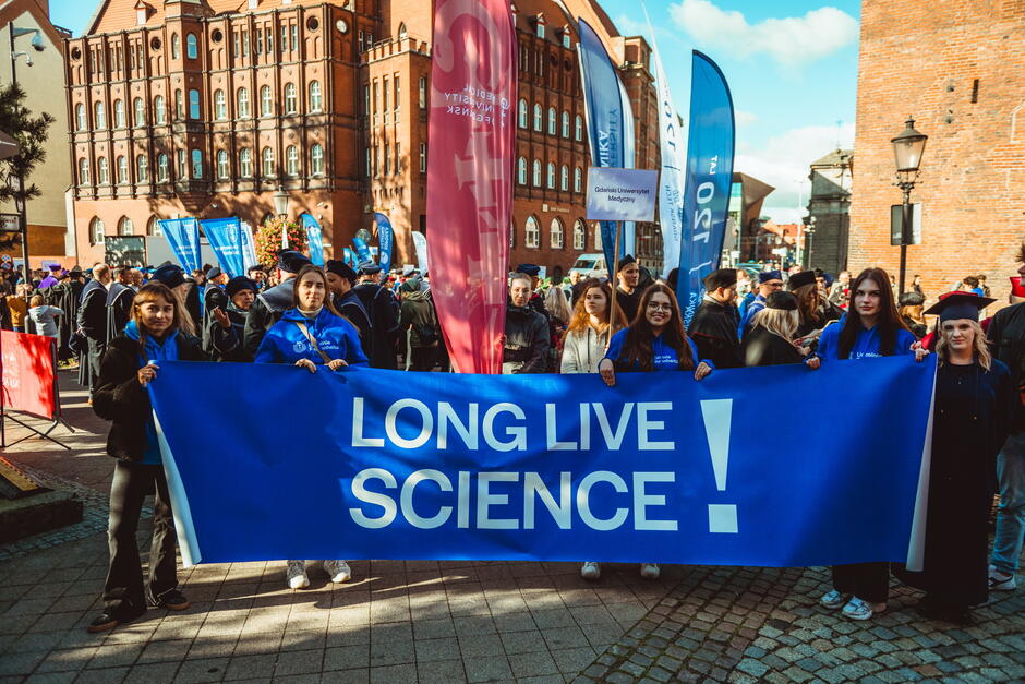 Na zdjęciu grupa studentów trzyma transparent z napisem „LONG LIVE SCIENCE!” podczas barwnego pochodu ulicznego w Gdańsku. W tle widać flagi uczelni, w tym Gdańskiego Uniwersytetu Medycznego, oraz liczne osoby uczestniczące w radosnym wydarzeniu akademickim.