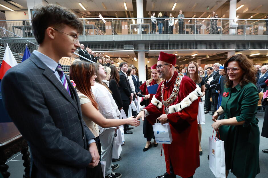 Na zdjęciu rektor ubrany w uroczysty strój akademicki wita się z nowymi studentami podczas oficjalnej inauguracji roku akademickiego. W tle widać uśmiechniętych gości oraz przedstawicieli uczelni, co podkreśla podniosły, ale też radosny charakter wydarzenia.