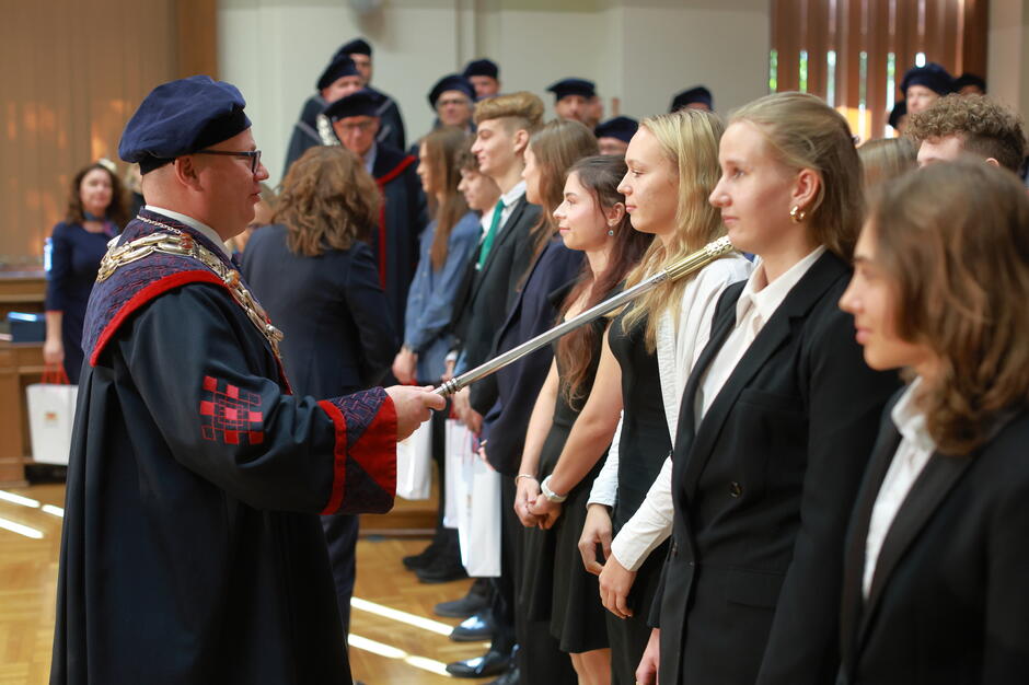 Na zdjęciu widać uroczystość immatrykulacji, podczas której rektor symbolicznie przyjmuje nowych studentów do społeczności akademickiej, dotykając ich ramienia berłem. W tle stoją profesorowie w togach, a nowo przyjęci studenci są odświętnie ubrani i ustawieni w szeregu.
