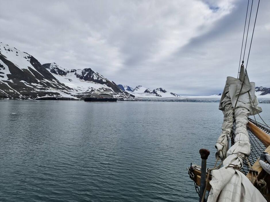 Na zdjęciu widać ośnieżone, surowe wybrzeże Arktyki z majestatycznymi górami i lodowcem spływającym do morza. Na wodzie unosi się duży statek pasażerski, a na pierwszym planie widoczny jest dziób żaglowca z opuszczonymi żaglami.
