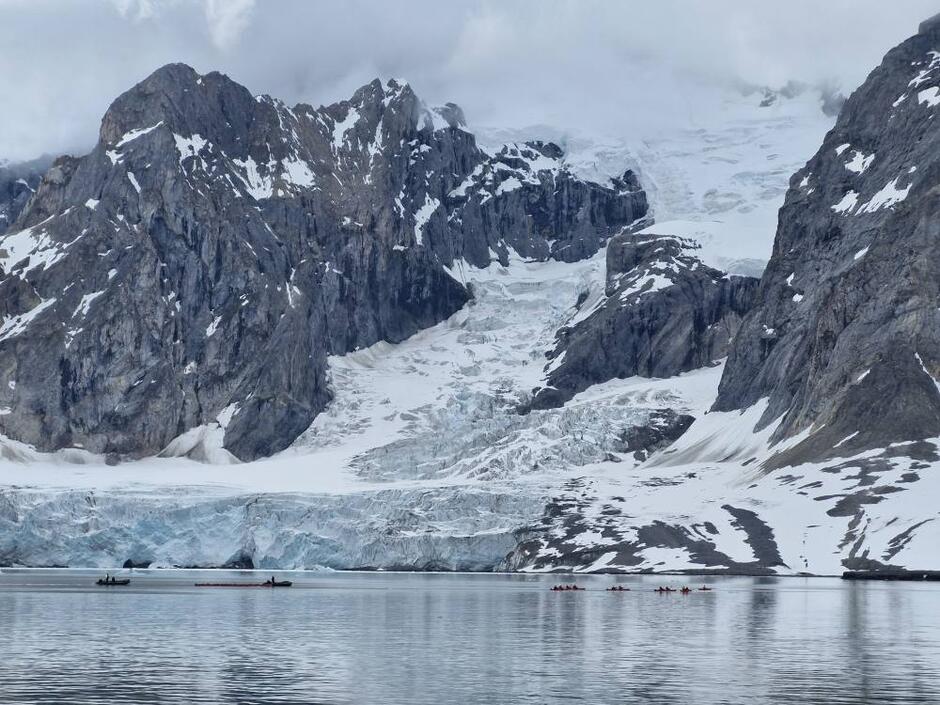 Na zdjęciu widać majestatyczne, skaliste góry pokryte śniegiem i lodowcem, schodzącym aż do spokojnej tafli wody. Na pierwszym planie płyną małe łodzie i kajaki, podkreślając ogrom przyrody i surowość arktycznego krajobrazu.
