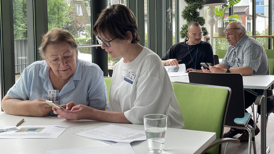 Na zdjęciu widać dwie pary osób siedzących przy stolikach w jasnym, nowoczesnym wnętrzu z dużymi oknami. Na pierwszym planie po lewej stronie starsza kobieta w jasnoniebieskiej koszuli patrzy na telefon, który trzyma kobieta obok niej — młodsza, w białej bluzce i okularach. Obie wydają się być zaangażowane w rozmowę lub naukę. Na stole leżą dokumenty i szklanka wody.  W tle po prawej stronie widać drugą parę — starszy mężczyzna z siwymi włosami i w okularach oraz młodszy mężczyzna w czarnej koszulce. Oni również wspólnie spoglądają na telefon.