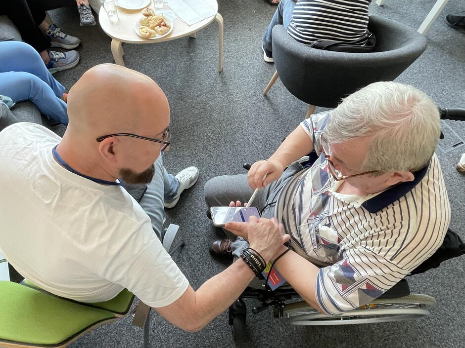 Na tym zdjęciu widać dwóch mężczyzn w trakcie rozmowy lub wspólnej pracy nad smartfonem. Starszy mężczyzna z siwymi włosami i okularami siedzi na wózku inwalidzkim i trzyma telefon, na którym coś pokazuje. Ma na sobie koszulkę w paski. Obok niego siedzi młodszy, łysy mężczyzna z brodą i okularami, ubrany w białą koszulkę. Widać, że obserwuje telefon z zainteresowaniem. W tle stoją fotele i niski stolik z ciastkami i szklankami, co sugeruje swobodną, warsztatową atmosferę. Zdjęcie prawdopodobnie przedstawia sytuację, w której młodszy człowiek pomaga starszemu w obsłudze smartfona lub rozwiązywaniu jakiegoś problemu technologicznego.