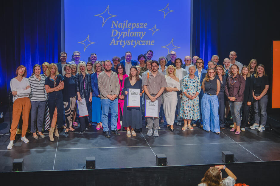 Na zdjęciu grupa osób stoi razem na scenie podczas uroczystości, na tle niebieskiej planszy z napisem „Najlepsze Dyplomy Artystyczne”. W centrum znajdują się laureaci trzymający dyplomy, otoczeni przez organizatorów i uczestników wydarzenia