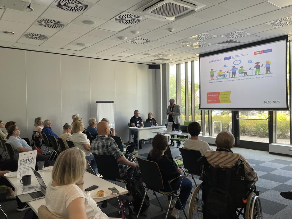 Na zdjęciu widzimy salę konferencyjną, w której odbywa się prezentacja lub warsztat. Po lewej stronie siedzi kilkanaście osób — głównie seniorów — uważnie słuchających prelegenta. Część z nich korzysta z wózków inwalidzkich. Przed nimi stoi ekran z prezentacją, na której widać logotypy projektów europejskich i hasło dotyczące włączenia cyfrowego: „Digital inclusion – leaving no-one behind in a digital world” Na ekranie widać również datę wydarzenia: 26.06.2025, oraz informację, że odbywa się ono w Gdańskim Centrum Kontaktów. Przy stole na środku siedzą dwie osoby, a jeden z mężczyzn stoi i przemawia. Zdjęcie przedstawia formalne spotkanie, prawdopodobnie w ramach projektu edukacyjnego dla seniorów lub osób z niepełnosprawnościami, mającego na celu wspieranie ich kompetencji cyfrowych i integracji społecznej.