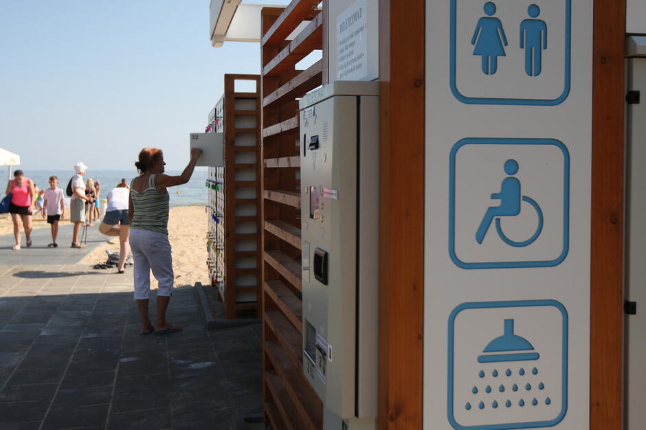 Na zdjęciu widzimy teren przy plaży z szafkami depozytowymi oraz automatami biletowymi, obok których znajdują się oznaczenia toalety, prysznica i udogodnień dla osób niepełnosprawnych. W tle spacerują ludzie, a całość znajduje się blisko morza. 