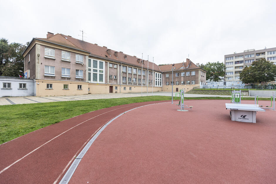 na zdjęciu fragment czerwonej bieżni, w tle budynek szkoły