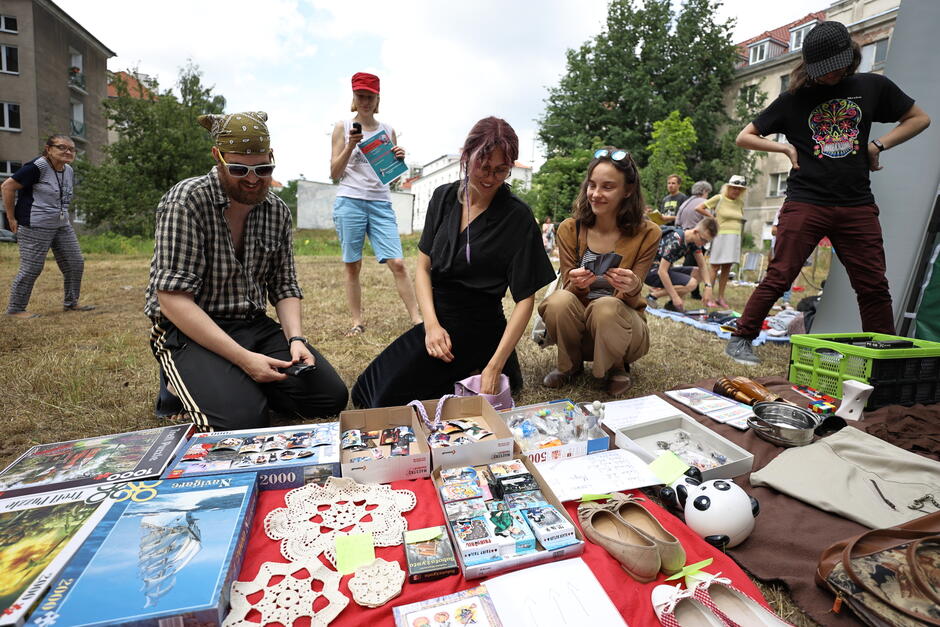 trzy dorosłe osoby kucają przy kocyku na którym widać między innymi naczynia i opakowania po puzzlach