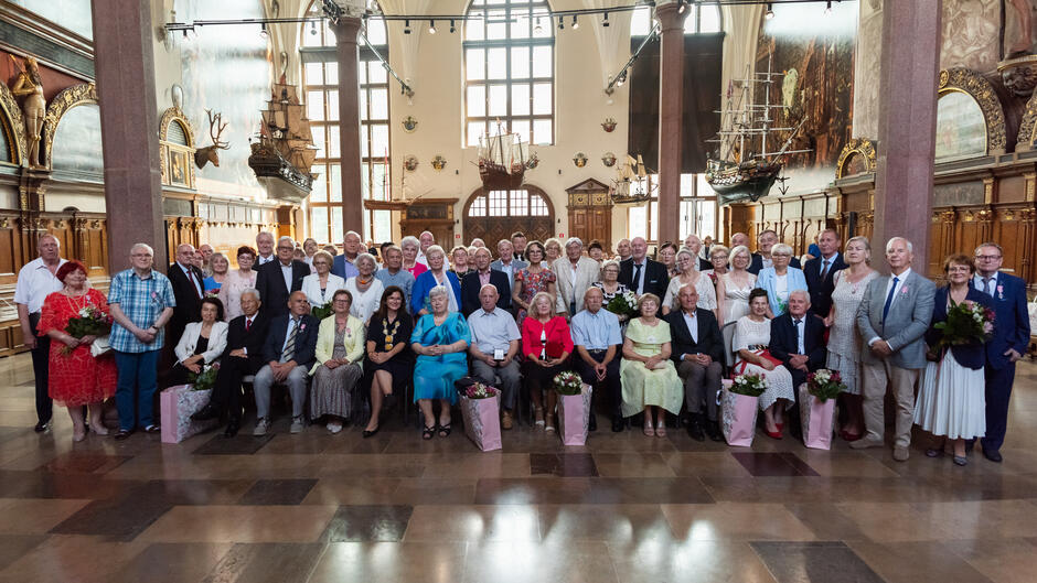 Na zdjęciu znajduje się duża grupa starszych osób, ustawionych do wspólnej fotografii w reprezentacyjnej sali z zabytkowym wystrojem i modelami żaglowców na ścianach. Wydarzenie wygląda na uroczystość jubileuszową, prawdopodobnie z okazji rocznicy małżeństwa, sądząc po odświętnych strojach i kwiatach w rękach uczestników