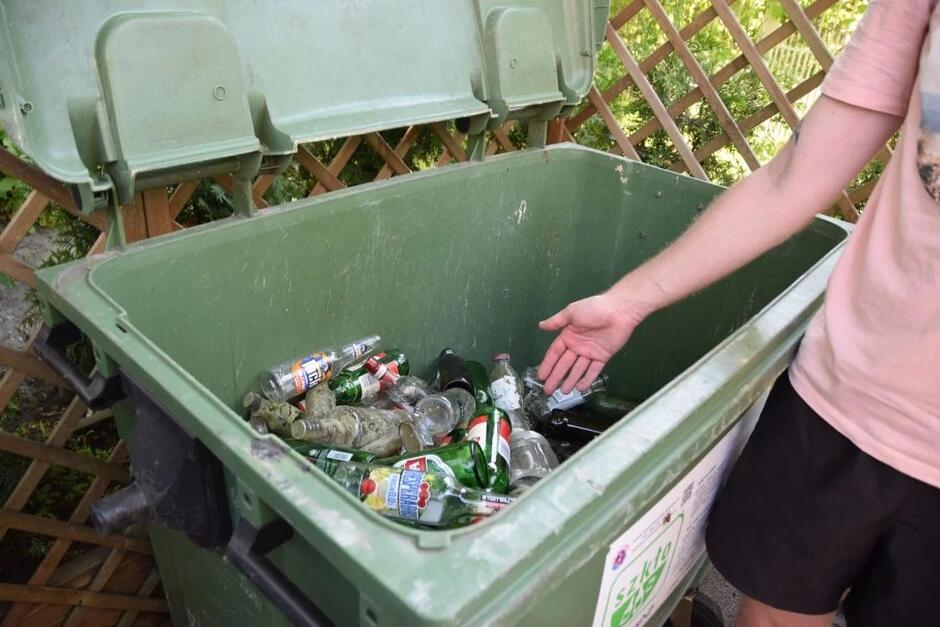 Ręka wskazuje na pojemnik na śmieci, w którym widać butelki.