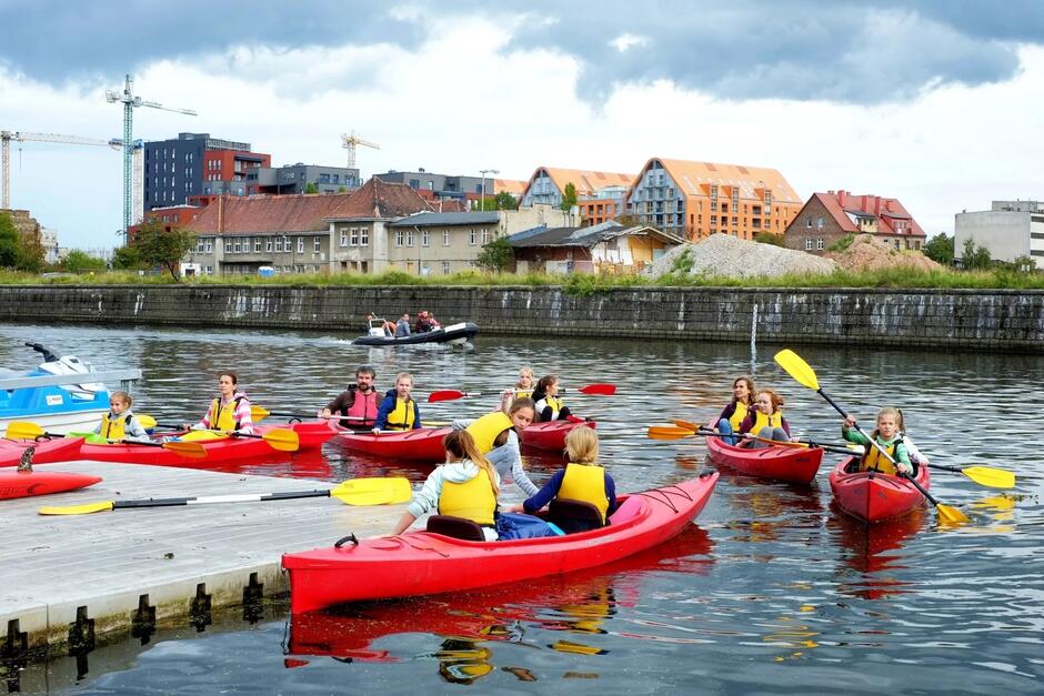 Grupa ludzi siedzi w kajakach na rzece w mieście.